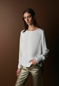 Valkoinen pitkähiha blouse pyöreällä kaula-aukolla ja rypytetyillä hihoilla, yhdistettynä kiiltäviin kultaisiin slim-fit-housuihin. Suora rakenne, minimalistinen muotoilu.