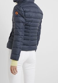 Veste matelassée bleu marine avec matelassage horizontal, col montant et patch logo orange sur l'épaule ; poignets côtelés jaunes ; matériau léger.