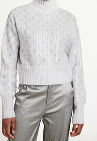 Pull gris clair en tricot avec un col montant côtelé, orné de sequins argentés, et des poignets côtelés, associé à un pantalon argenté brillant.