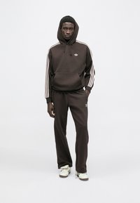 Camisola desportiva castanha com capuz e bolso frontal, apresentando três riscas brancas em cada manga. Combinada com calças de treino castanhas a condizer e sapatos brancos.