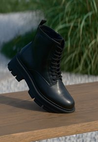 Bottines en cuir noir à lacets avec une semelle épaisse et un orteil renforcé. Présente une finition lisse et des détails de couture le long de la semelle.