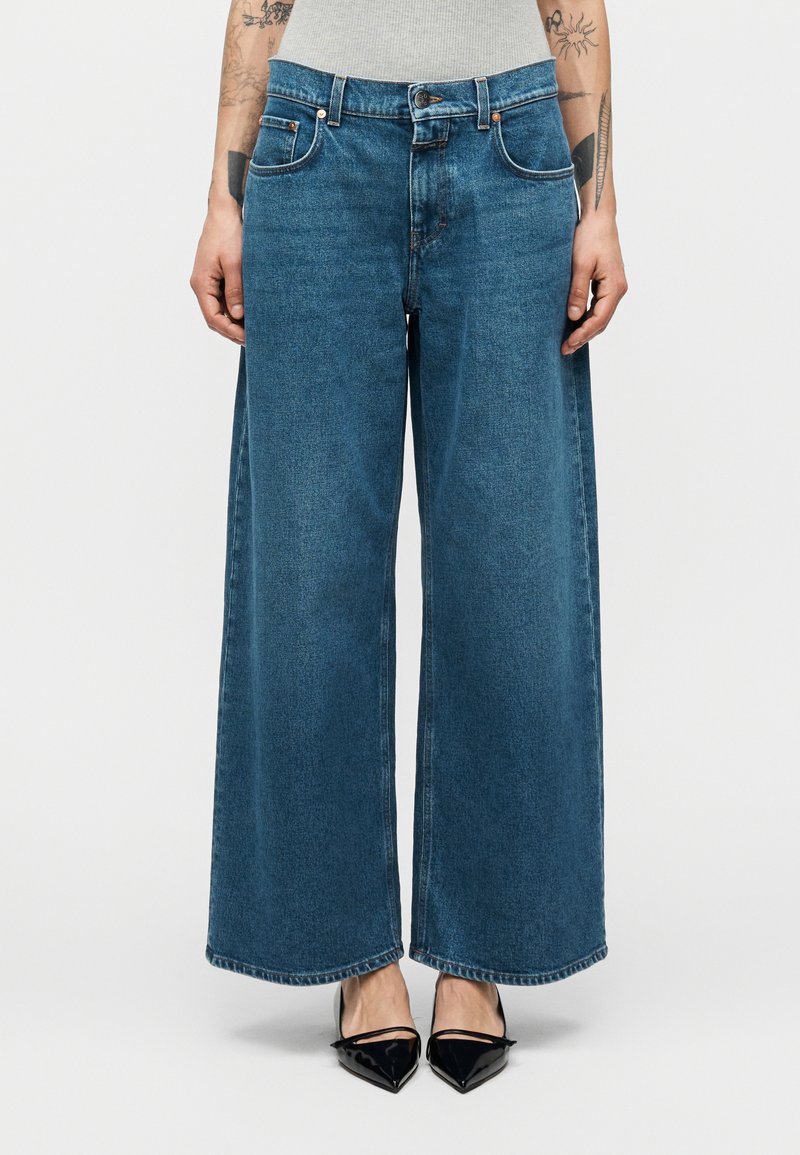 Τζιν παντελόνι με φαρδιά μπατζάκια σε μπλε αποχρώσεις, ψηλή μέση, σχέδιο με πέντε τσέπες και αντίθετα ραφές. Συνδυασμένο με μαύρες γόβες με μυτερό πάτο.