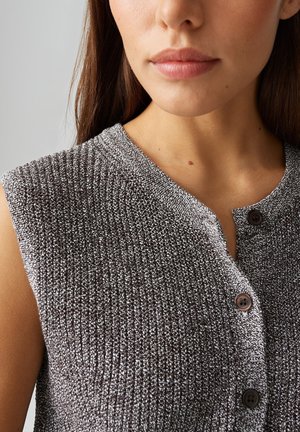 Jeune femme portant un haut sans manches en maille grise texturée avec des boutons noirs sur le devant, montrant son cou et une partie de son visage.