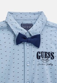 Chemise bleu clair avec col pointu, nœud papillon bleu marine et logo brodé noir. Petits motifs floraux noirs et blancs sur l'ensemble.