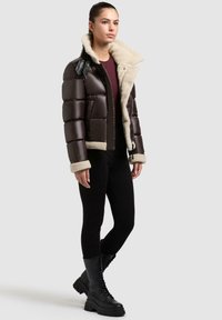 Braune gepolsterte Jacke mit cremefarbigem Kunst-Lammfellkragen und -futter, schwarzen Kunstleder-Akzenten, Reißverschluss und elastischen Bündchen. Schwarze Leggings und Stiefel.