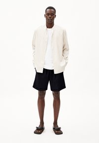 Gestreept beige overhemd met knopen, wit T-shirt, zwarte shorts en zwarte sandalen. Het overhemd is lichtgewicht; de shorts zijn knielang met een elastische taille.