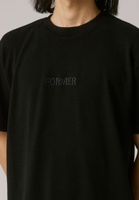 Black cotton t-shirt with short sleeves, featuring an embroidered "FORMER" logo in darker thread on the chest. Textured fabric.