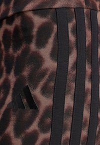 Κοντινή λήψη υφάσματος με καφέ και μαύρο λεοπάρ μοτίβο, με τρεις κάθετες μαύρες ρίγες και ένα μαύρο κεντημένο λογότυπο Adidas.