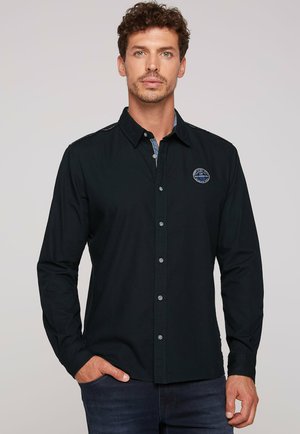 Black long-sleeve shirt with button closure, featuring a textured fabric and a logo patch on the left chest. Accents with contrasting blue inside collar.