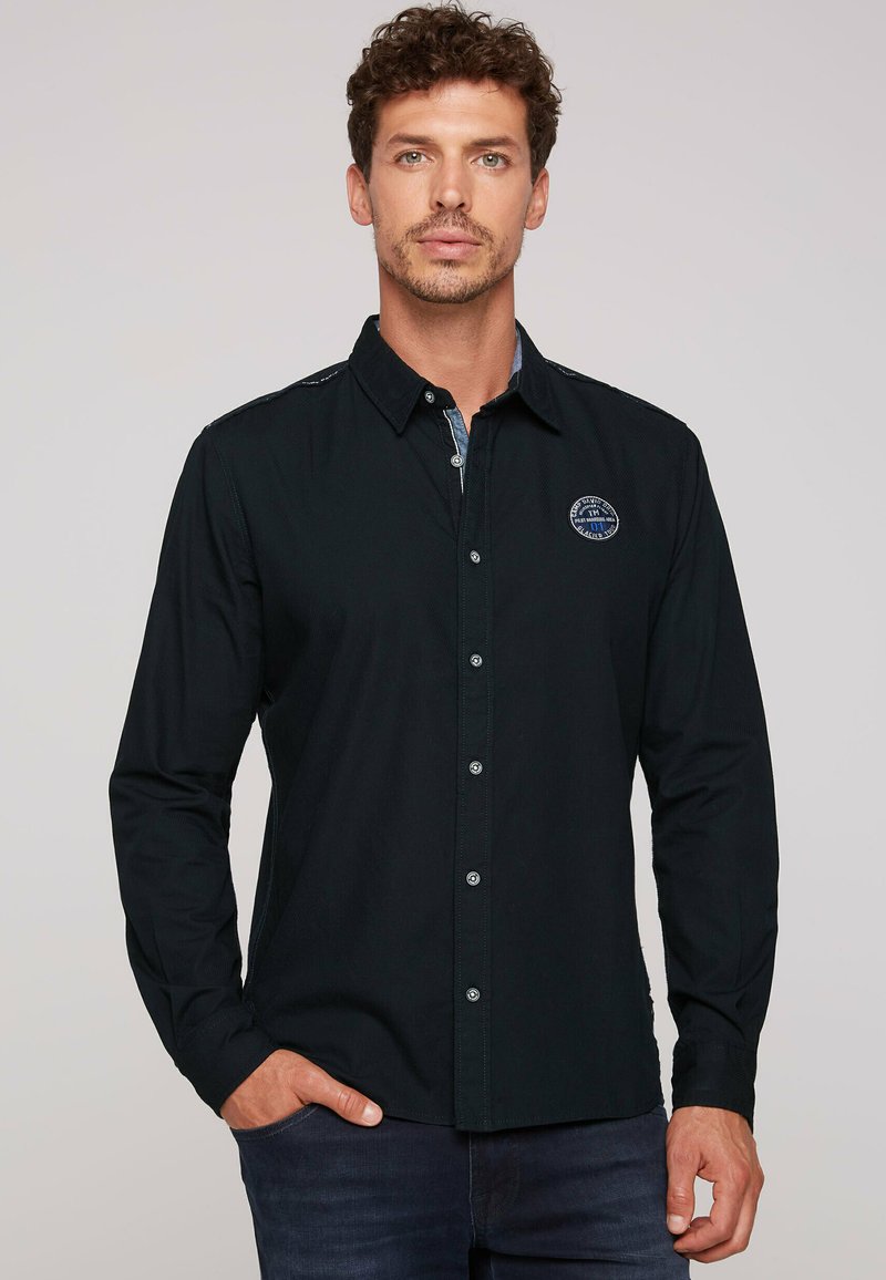 Camisa negra de manga larga con cierre de botones, con un tejido texturizado y un parche del logo en el lado izquierdo del pecho. Detalles con un color azul contrastante en el interior del collar.