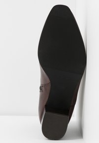 Chaussure en cuir marron avec un bout pointu, dessus texturé et une semelle en caoutchouc noir avec des rainures horizontales pour la traction.