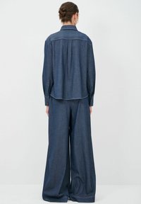Completo con camicia in denim e pantaloni a gamba larga di colore blu scuro. Presenta un colletto, polsini con bottoni e cuciture visibili lungo i bordi.