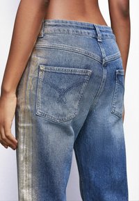 Jeans de denim con un lavado azul desgastado. Presentan dos bolsillos traseros con costuras decorativas y acentos de hardware dorado a lo largo de los laterales.