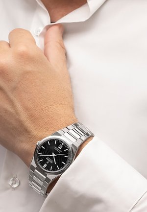 Linkes Handgelenk mit silberner Metalluhr mit schwarzem Zifferblatt und Datumsanzeige, getragen mit einem weißen Langarmhemd.