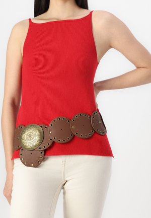Femme portant un top rouge sans manches et une large ceinture marron avec de gros clous ronds et une boucle décorative en laiton, associée à un pantalon clair.