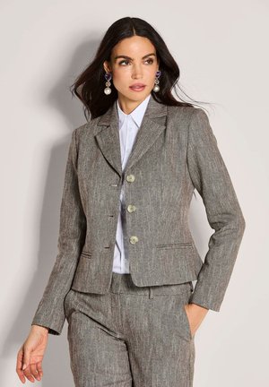 Vrouw in een grijze herringbone kostuum met een wit shirt, met grote parel en paarse hanger oorbellen, poserend tegen een neutrale achtergrond.