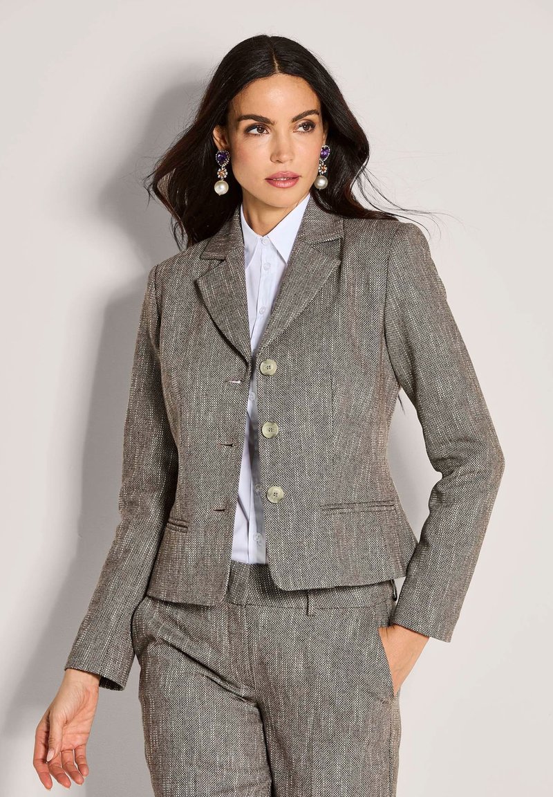 Vrouw in een grijze herringbone kostuum met een wit shirt, met grote parel en paarse hanger oorbellen, poserend tegen een neutrale achtergrond.