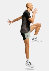 Persona atlética con camiseta negra, pantalones cortos bicolores y zapatillas de running blancas posando en medio de un paso frente a un fondo simple.