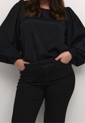 Woman wearing a black satin long-sleeve blouse with puffed sleeves and black fitted pants, hands tucked into front pockets.