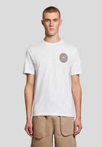 Νέος άνδρας που στέκεται, φοράει λευκό κοντομάνικο t-shirt με κυκλικό λογότυπο στο στήθος και μπεζ cargo σορτς, μπροστά σε απλό φόντο.