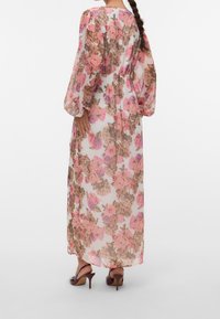 Femme aux cheveux foncés tressés portant une longue robe fleurie transparente avec des fleurs roses et marron et des manches ballon transparentes, associée à des sandales marron à talons.