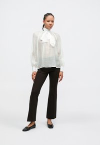 Λευκό, διαφανές πουκάμισο με λεπτομέρεια δέσιμου στο λαιμό· μακριά, φουσκωτά μανίκια. Συνδυάζεται με εφαρμοστά σκουρόκαφε παντελόνια και μαύρα φλατ παπούτσια. Απλός σχεδιασμός.