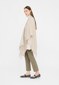 Poncho beige con frange, texture morbida, design drappeggiato sopra una maglietta bianca, abbinato a pantaloni verde oliva e scarpe slip-on nere con dettagli.