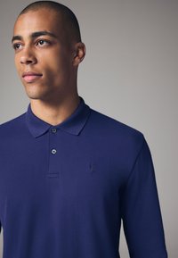 Mornarska polo majica z dolgimi rokavi, izdelana iz gladkega bombaža. Ima klasičen ovratnik, zapenjanje na dva gumbka in vezeno logo na prsih.