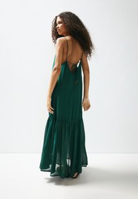 Rochie maxi verde, cu bretele subțiri, spate deschis cu detaliu de legătură și fustă fluidă în straturi, realizată dintr-un material ușor.