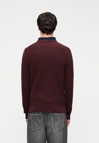 Pull en maille bordeaux avec un col marine, des manches longues et une texture côtelée, associé à un jean gris, vu de dos.