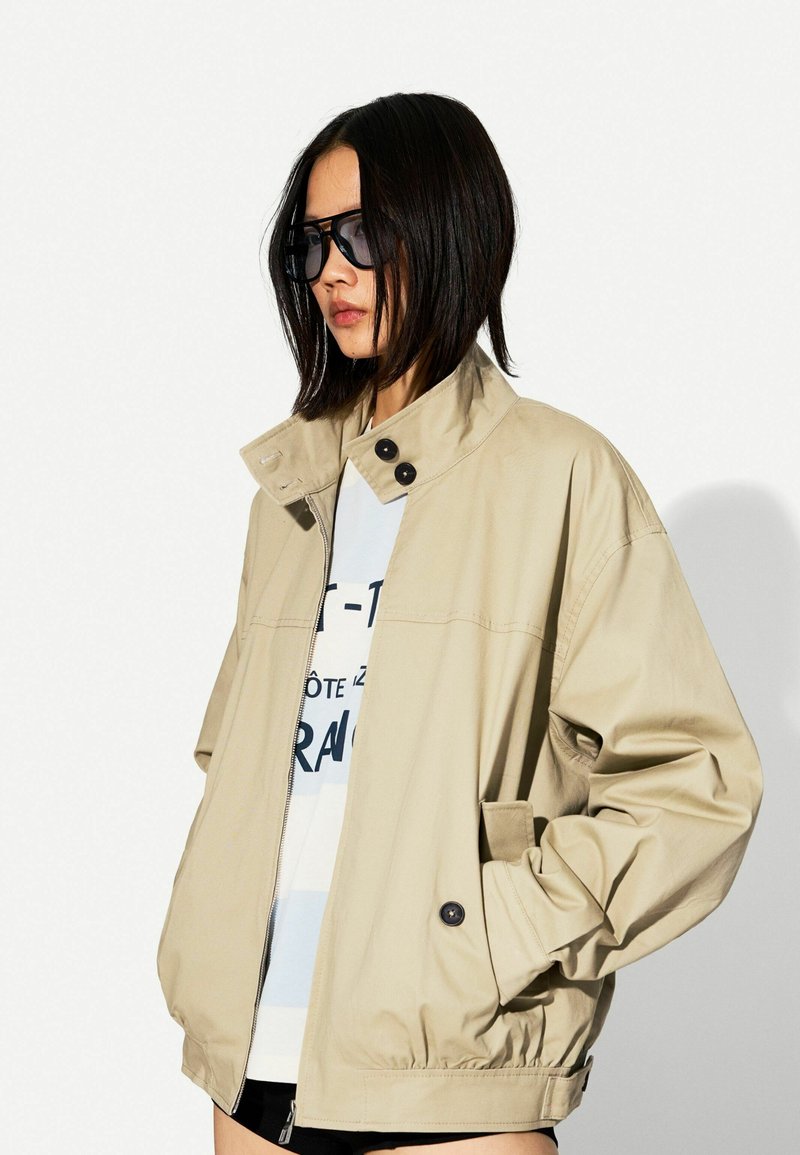 Beige, leichte Jacke mit Kragen, Reißverschluss vorne und Druckknöpfen. Verfügt über seitliche Taschen und elastische Bündchen für eine taillierte Passform.