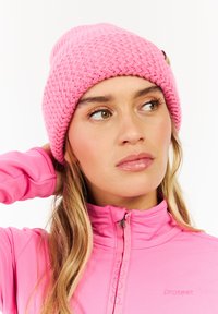 Pink knitted beanie with a textured pattern and a small logo detail. Worn with a matching pink zip-up top in a close-up view.