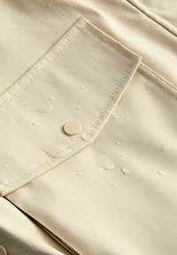 Bežová látka odolná voči poveternostným vplyvom s viditeľnou kapsou, ktorá má zaoblené gombíky a kvapky vody na povrchu, zvýrazňujúca textúru.