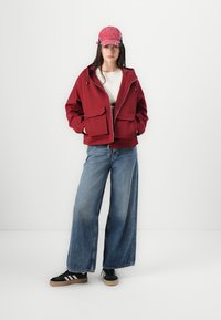 Burgunderfarget hettegenser med store front lommer, kombinert med en hvit skjorte og vide blå jeans. Modellen har på seg en rosa caps og svarte joggesko.