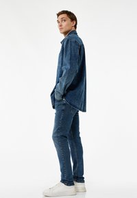 Chaqueta de denim azul con un corte holgado y bolsillos frontales, combinada con jeans azules ajustados y zapatillas blancas, todo sobre un fondo liso.