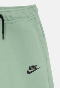Svetlozelené Nike tepláky s elastickým pásom, šnúrkou, vreckami a čiernym logom na ľavom stehne. Mäkká tkanina.
