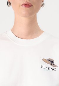 Hvit bomulls T-skjorte med brodert stråhatt og teksten "BI AZING" i svart og lyseblått. Enkel design med rund hals.