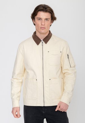 Man draagt een crème jas met een bruine corduroy kraag, rits aan de voorkant, borst- en zijzakken, staand tegen een effen lichte achtergrond.