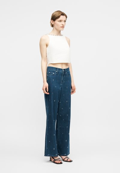 Femme debout portant un crop top blanc sans manches, un jean large bleu avec de petits motifs blancs, et des sandales noires à lanières.