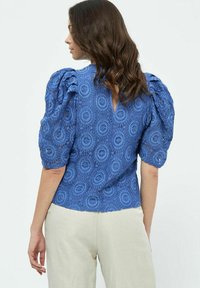 Blusa in pizzo blu con maniche a sbuffo, motivi di ricamo circolari e apertura a V sul retro. Tessuto texture e silhouette aderente.