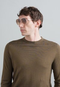 Camicia a maniche lunghe marrone indossata da una persona con occhiali da sole tondi e colorati con una montatura argentata. Texture liscia e design aderente.