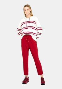 Pullover bianco con strisce orizzontali rosse e logo, abbinato a pantaloni sportivi rossi a gamba affusolata e sneakers rosse. L'outfit presenta una vestibilità rilassata.