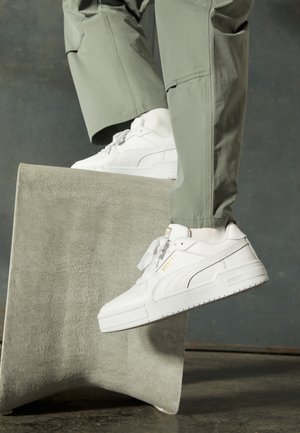 Baskets blanches en cuir lisse, avec un bout arrondi et une semelle plate en caoutchouc. Détail du logo doré sur le côté et lacets noués. Design décontracté.