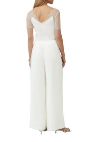 Débardeur en dentelle blanche à manches courtes, décolleté dans le dos, associé à un pantalon large blanc. Texture douce, tombé élégant, design minimal.