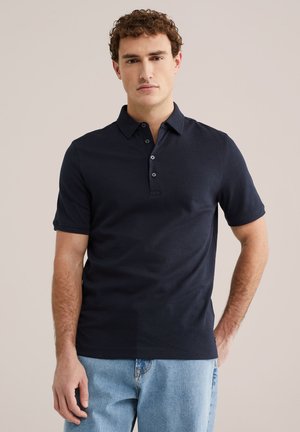 Fiatal férfi, fekete, háromgombos, testhezálló galléros pólót és világoskék farmert viselve, egyszínű bézs háttér előtt állva.