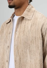 Beige corduroy jasje met een geribbelde textuur, voorzien van een klassieke kraag en knopen aan de voorkant. Draagt losjes over een wit shirt en heeft een relaxte pasvorm.