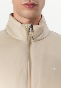 Beige Daunenjacke mit hohem Kragen, Frontreißverschluss und Logodetail. Glatte Textur; leichtes Design, das sich gut zum Layering eignet.