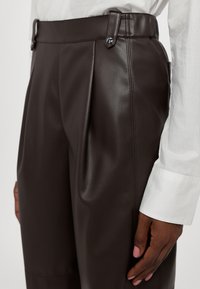 Pantalon en cuir marron foncé avec plis sur le devant et passants de ceinture, porté avec une chemise blanche à manches longues et une main avec un vernis à ongles rose visible.