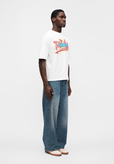 Λευκό κοντομάνικο μπλουζάκι με πολύχρωμο γραφικό σχέδιο, συνδυασμένο με ανοιχτό μπλε τζιν παντελόνι φαρδιάς γραμμής και μπεζ παπούτσια τύπου slip-on.