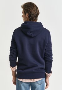 Felpa blu navy con design rilassato, dotata di cappuccio ampio, polsini e orlo a costine, indossata sopra una maglietta a righe rosa, vista da dietro.